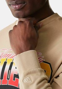 Lähenemine inimese käele, mis on lähedal lõuale, kandes beežikat sviitrit, millel on multivärviline graafiline tekst rinnal ja soonikkoes detaalid varrukatel.