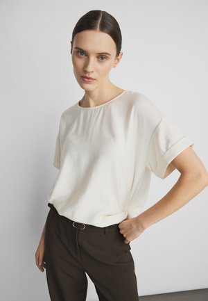 Kvinde med mørkt hår trukket tilbage står mod en ensfarvet baggrund iført en løs hvid kortærmet top og mørke bukser med bælte.