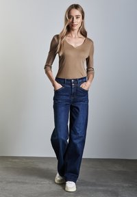 Maglietta aderente a maniche lunghe di colore beige con scollatura a V. Jeans a gamba larga di colore blu scuro con tasche. Modella in piedi con le mani in tasca, sfondo neutro.