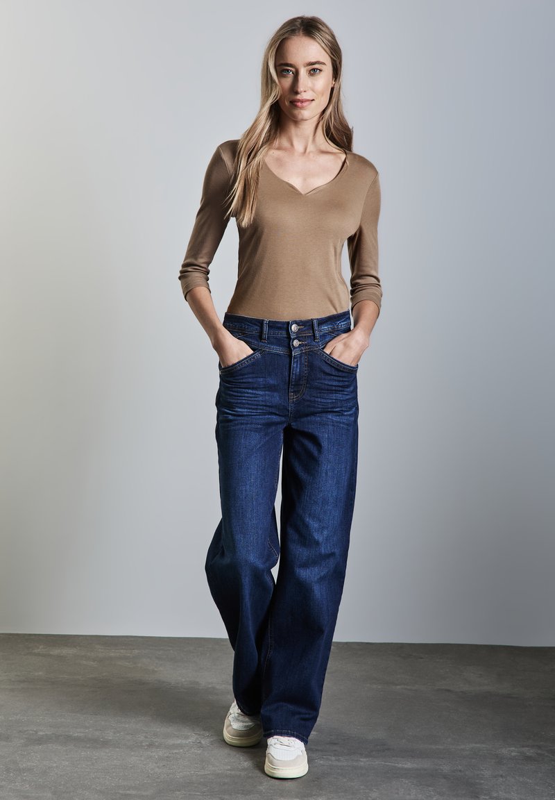 Maglietta aderente a maniche lunghe di colore beige con scollatura a V. Jeans a gamba larga di colore blu scuro con tasche. Modella in piedi con le mani in tasca, sfondo neutro.