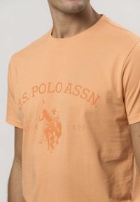 Mężczyzna w brzoskwiniowej koszulce z napisem U.S. Polo Assn. i grafiką przedstawiającą gracza polo na froncie.