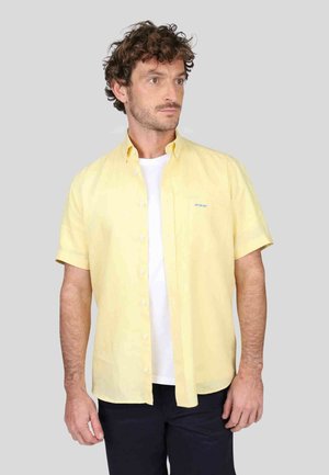 Chemise à manches courtes, boutonnée, en tissu jaune clair, avec une texture subtile et un petit logo bleu sur le côté gauche. Portée par-dessus une chemise blanche.