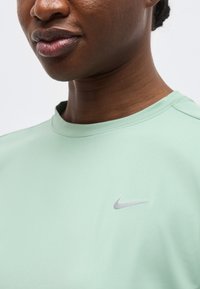 Κοντινό πλάνο ενός ατόμου που φοράει ένα ανοιχτό πράσινο πουκάμισο Nike με ένα μικρό ανακλαστικό λογότυπο Nike στο στήθος.