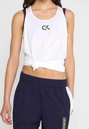 Top sin mangas blanco con un dobladillo anudado, que presenta un logo negro, combinado con joggers azul marino acentuados por paneles laterales blancos y una etiqueta de logo.