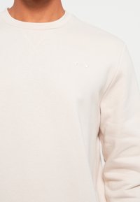 Sudadera de cuello redondo en beige claro, fabricada en una suave mezcla de algodón. Presenta un pequeño logotipo bordado en el pecho y un cuello acanalado.