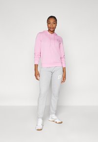 Rosa Kapuzenpullover mit einem Logo auf der Vorderseite, kombiniert mit grauen Jogginghosen mit einem weißen Grafikdesign, ergänzt durch weiße Sportschuhe.