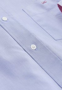 Chemise bleu clair à manches longues en tissu texturé, dotée de boutons blancs, d'une poche et d'un petit détail en forme de nœud rose.