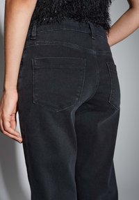 Vue arrière d'une personne portant un jean noir taille haute avec deux poches arrière et un haut sans manches noir texturé.
