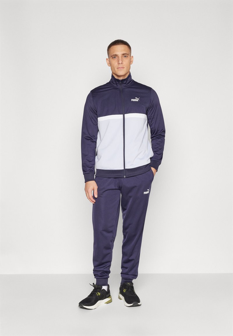 Dunkelblauer Puma-Trainingsanzug mit grauem Brustpanel, Reißverschlussjacke und passender Hose. Mit elastischen Bündchen und Logo-Akzenten. Schwarze Sportschuhe.