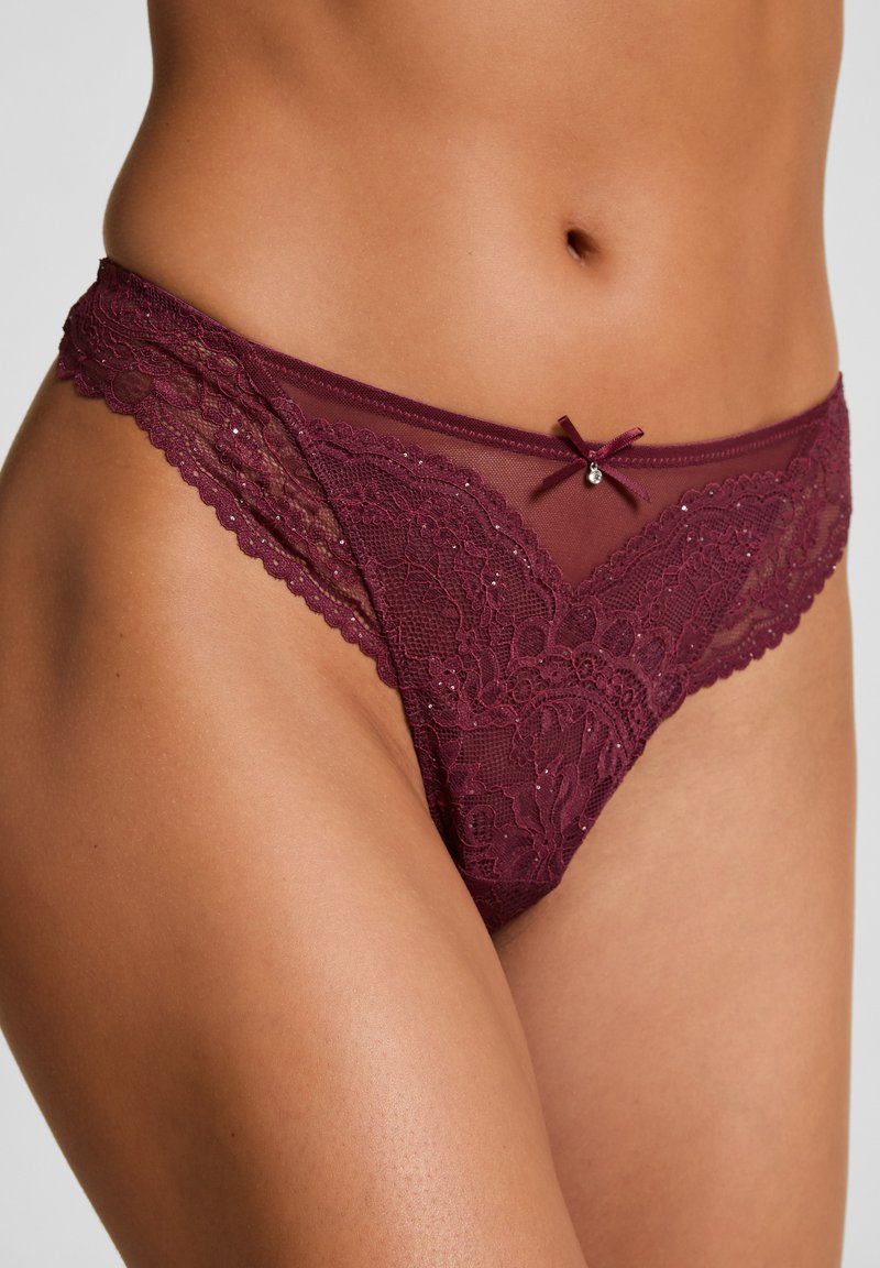 Vrouw in bourgondische kant bikini-stijl ondergoed met een klein strikje en juweel detail aan de voorkant van de tailleband.