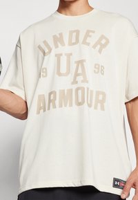 Ljusbeige oversized t-shirt med "UNDER ARMOUR" grafik i mörkare beige. Mjuk bomullstyg, korta ärmar, rundad halsringning och logotypmärke.