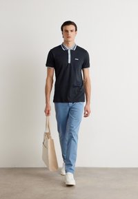 Mornarska polo majica s svetlo modrim prugastim ovratnikom, slim fit, v kombinaciji s svetlo modrimi hlačami in belimi supergami, nosi bež platneno torbo.