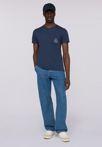 Camiseta de algodón azul marino con un bolsillo en el lado izquierdo del pecho y un gráfico de color claro, combinada con jeans de mezclilla azul claro y zapatillas blancas.