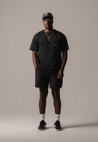 T-shirt noir avec un petit logo sur la poitrine, shorts noirs, chaussettes blanches et baskets noires. Porté avec une casquette grise et des colliers en or superposés.