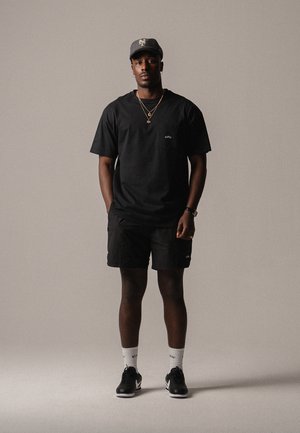 T-shirt noir avec un petit logo sur la poitrine, shorts noirs, chaussettes blanches et baskets noires. Porté avec une casquette grise et des colliers en or superposés.