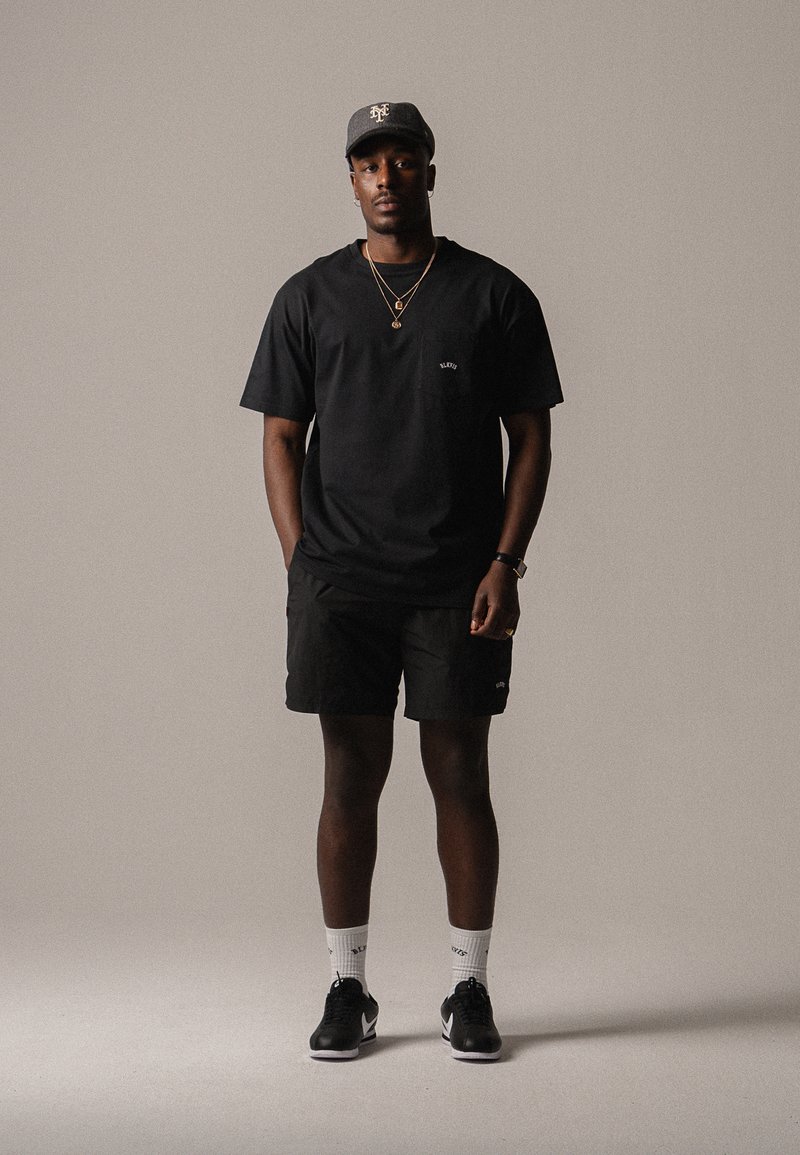 T-shirt noir avec un petit logo sur la poitrine, shorts noirs, chaussettes blanches et baskets noires. Porté avec une casquette grise et des colliers en or superposés.