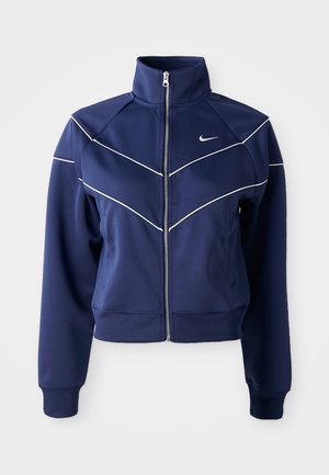 Marineblå zip-jakke laget av glatt stoff har høy krage, hvit chevron-søm og sidelommer. Nike-logo på brystet.