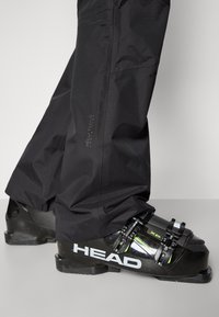 Botas de esqui pretas com logo "HEAD" branco, fivelas ajustáveis e acabamentos verdes. Combinadas com calças impermeáveis pretas feitas de tecido texturizado.