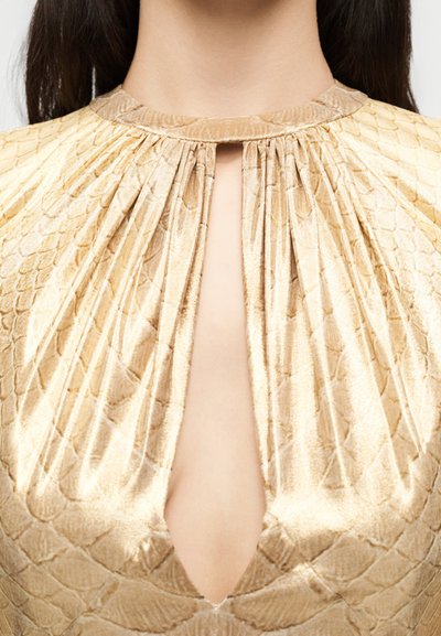 Κοντινό πλάνο μπεζ μπλούζας με σχέδια, υφασμένη υφή και λαιμόκοψη με άνοιγμα που δείχνει το πάνω μέρος του στήθους και του λαιμού.