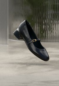 Μαύρο δερμάτινο παπούτσι Mary Jane με μικρό τακούνι και χρυσή πόρπη, εμφανιζόμενο να αιωρείται πάνω από γκρίζο τσιμεντένιο πάτωμα μπροστά σε φόντο υφασμένο γυαλί.