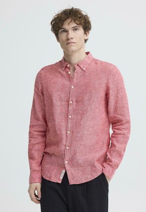 Young man with curly hair wearing a long-sleeve red linen button-up shirt and black pants, standing with one hand in pocket.