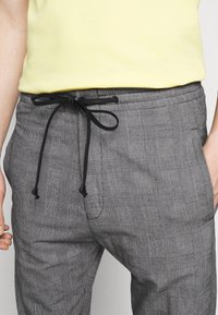Szare spodnie w kratę wykonane z lekkiego materiału, z czarnym ściągaczem w pasie i bocznymi kieszeniami. Wzór obejmuje cienkie linie i kwadraty.