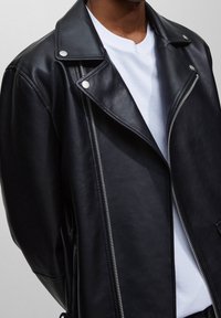 Chaqueta de motociclista de cuero negro con herrajes plateados, cierre frontal con cremallera y textura suave. Llevada sobre una camiseta blanca.