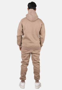 Sudadera con capucha beige y pantalones joggers a juego, que presentan un material suave, puños acanalados y un ajuste relajado. Las zapatillas blancas completan el conjunto.