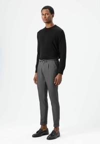 Schwarzer Pullover mit langen Ärmeln, graue plissierte Hose und schwarze Loafers. Die Hose ist maßgeschneidert mit umgeschlagenem Saum. Glatte Texturen überall.