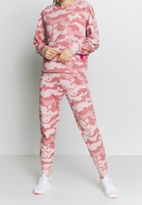 Różowa bluza z kapturem i dopasowane spodnie w kamuflażowym wzorze, wykonane z miękkiego materiału, o swobodnym kroju i ściągaczach przy rękawach, w połączeniu z białymi trampkami.