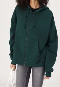 Sudadera con cremallera de gran tamaño en color verde oscuro, con capucha ajustable y bolsillos frontales, fabricada en tejido suave, con puños y dobladillo acanalados.