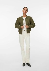 Femme portant une veste bomber boutonnée vert olive, un haut blanc, un pantalon blanc et des bottes noires pointues, debout devant un fond blanc uni.