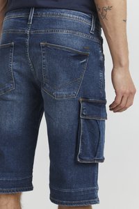 Tamno plave traper kratke hlače s velikim bočnim džepovima i stražnjim džepovima, koje nosi osoba s vidljivim tetovažama na podlaktici.