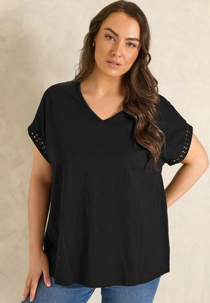 Kobieta z długimi brązowymi włosami, ubrana w luźną czarną bluzkę z dekoltem w kształcie litery V z wzorzystymi krótkimi rękawami i niebieskie dżinsy, stojąca na jednolitym tle.