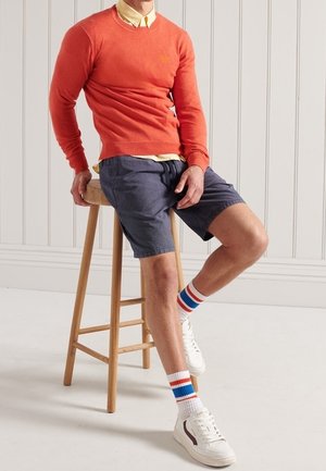 Pull rouge-orange, chemise jaune à col, short bleu marine. Baskets blanches avec des accents marron et chaussettes rayées. Personne assise sur un tabouret en bois.