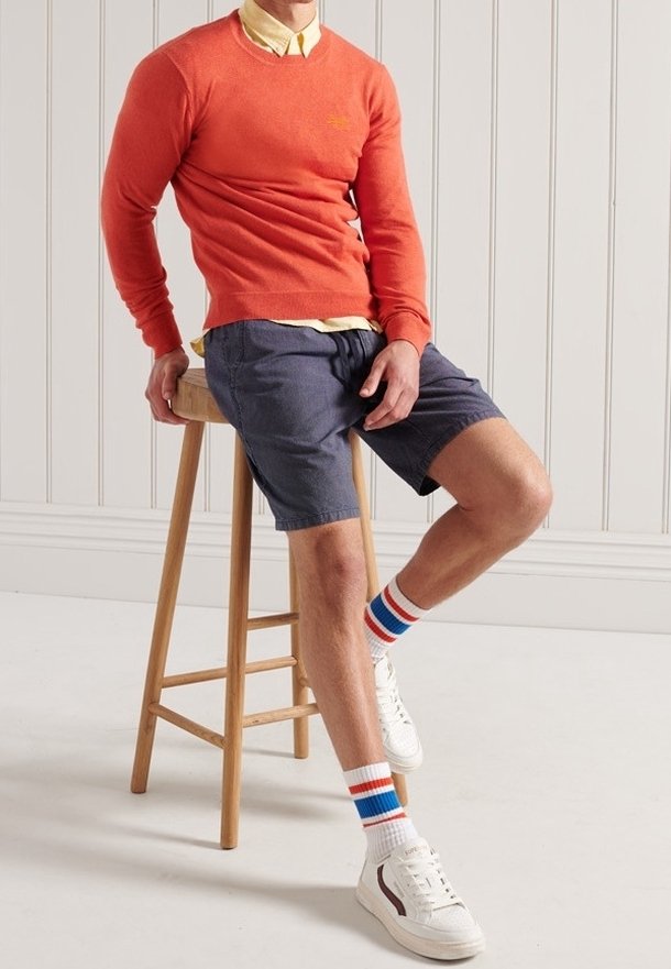 Pull rouge-orange, chemise jaune à col, short bleu marine. Baskets blanches avec des accents marron et chaussettes rayées. Personne assise sur un tabouret en bois.
