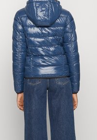 Marineblaue Puffjacke mit glänzender Oberfläche, horizontalem Steppmuster, abnehmbarer Kapuze und schwarzem Saum. Kombiniert mit weit geschnittenen Jeanshosen.