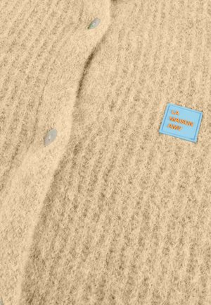 Pull en maille beige avec un motif texturé, doté de boutons ronds et clairs ainsi que d'une étiquette bleue portant l'inscription "LA MAISON AMV" en lettres orange.