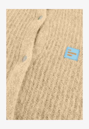 Beežist kootud kampsun tekstuurse mustriga, millel on ümmargused heledad nupud ja sinine silt oranži kirjaga "LA MAISON AMV".