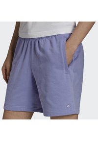 Osoba v světlých fialových volnočasových šortkách s elastickým pasem a bočními kapsami, jedna ruka v kapse, malé logo blízko lemu.