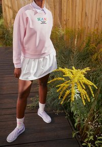 Sudadera rosa con el logo "KANI", falda blanca de volantes, zapatillas de cuadros morados y calcetines blancos, todo con un fondo de madera.