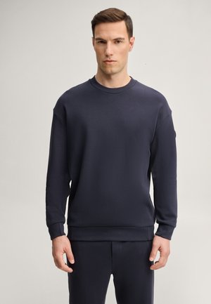 Uomo con capelli castani corti sta in piedi rivolto in avanti, indossa una semplice felpa blu scuro e pantaloni abbinati su uno sfondo chiaro.