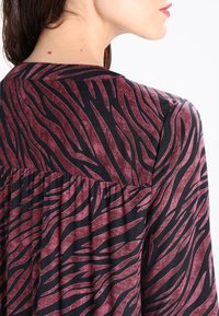 Tissu noir avec un motif de rayures zèbre bordeaux ; design plissé à l'arrière ; texture douce, col rond et coupe décontractée.