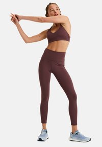 Un crop top de sport bordeaux et un legging taille haute en tissu lisse et extensible. Le modèle effectue une pose d'étirement, portant des chaussures de sport grises.