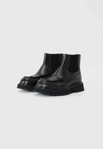 Bottines en cuir noir avec semelle en caoutchouc, panneaux élastiques sur les côtés, détails texturés et un design distinctif avec une large sangle sur le cou-de-pied.