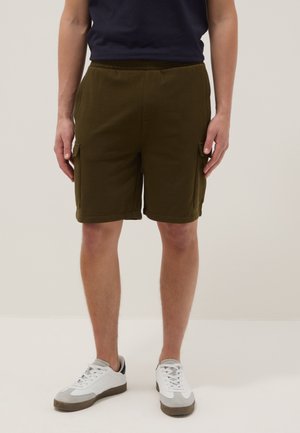 Mann trägt dunkelolivfarbene Cargo-Shorts, weiße und graue Sneakers, steht vor einem einfarbigen hellen Hintergrund.