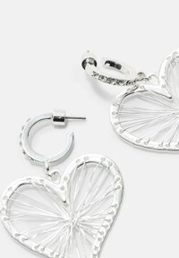 Orecchini a cerchio in argento con pendenti a forma di cuore a giorno, caratterizzati da dettagli in filo di metallo che si irradiano e piccoli cristalli incastonati.