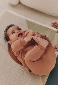 Pehme, roostevärvi beebiriietus elastsete mansettidega. Beebi lebab tekstuuriga matil ja naeratab, hoides kätega oma jalgu.