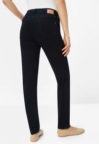 Donkerblauwe broek met een getailleerd ontwerp, achterzakken en een leren label op de tailleband. Gladde textuur met een lichte glans.