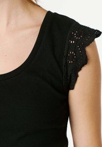 Κοντινό πλάνο ενός ατόμου που φοράει μαύρο πλεκτό κορμάκι με κοντά μανίκια από δαντέλα, διακοσμημένα με λουλουδάτα μοτίβα κοψίματος.